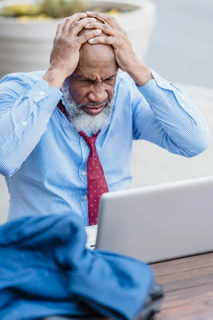 A man in a blue dress shirt and red tie is shown in front of a computer with his hands to his head and eyes closed in frustration.