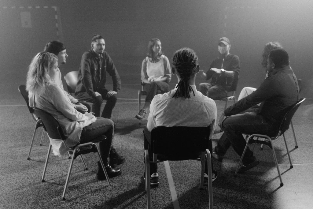 A black and white photo of a group of people sitting in chairs, all facing each other in a circle.