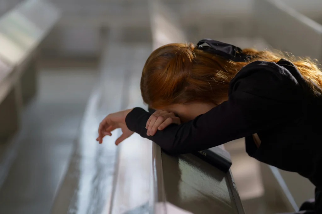 A woman in black is sitting in a church pew, head lowered against the pew in front of her, presumably crying