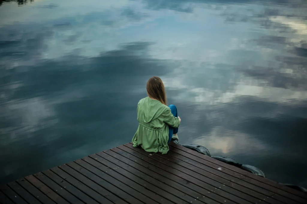 A woman is sitting on the edge of a dock, facing the water. You can see her back and the reflection of the clouds in the water.